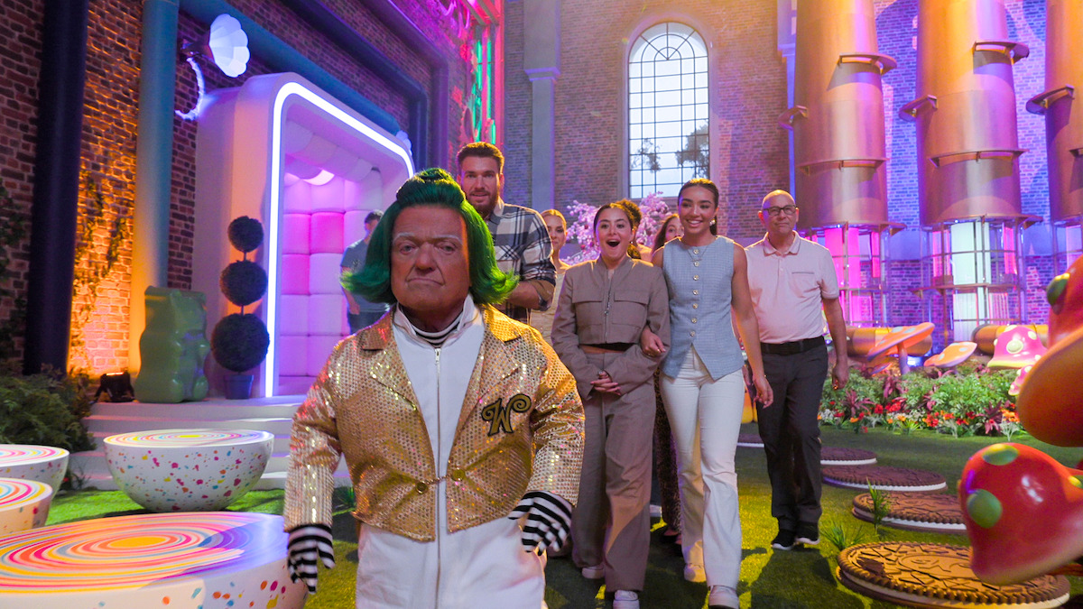 A person in a gold jacket and green wig leads a group of adults through a colorful, whimsical candy-themed set with bright lights, large windows, and oversized sweets.