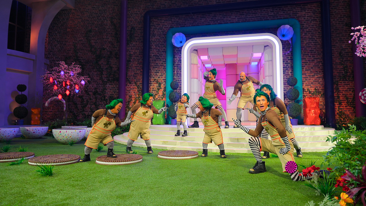 Group of performers dressed as Oompa Loompas with green hair and brown outfits dance on a vibrant, whimsical set with artificial grass, colorful lights, and a futuristic doorway, evoking a playful, fantasy factory environment.