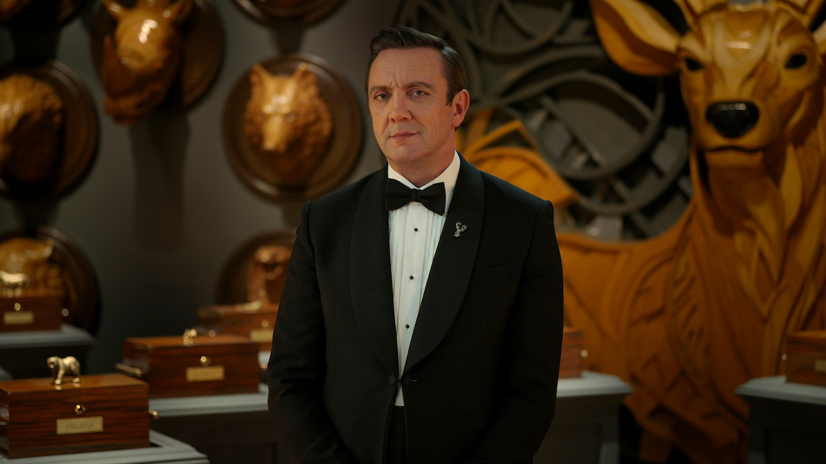 Man in a black tuxedo stands in a room decorated with large carved wooden animal heads and boxes, giving a formal and elegant atmosphere.