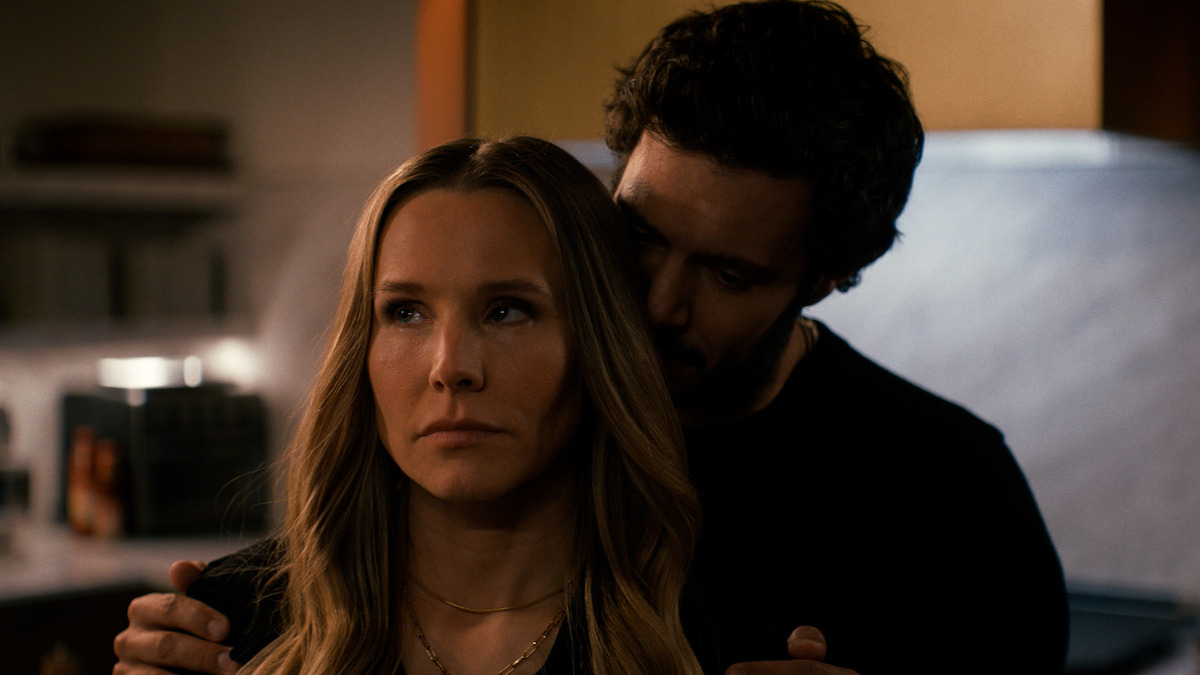 Adam Brody as Noah embracing Kristen Bell as Joanne from behind in a dimly lit room. 