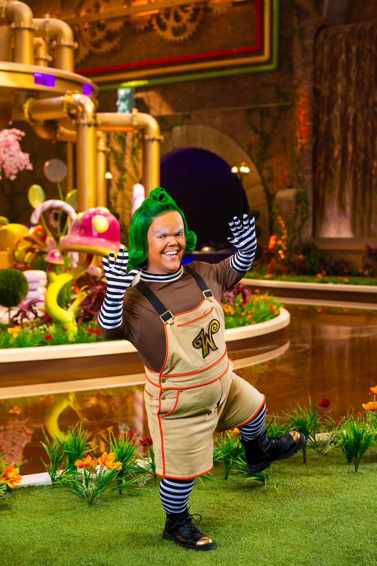 Person dressed as an Oompa Loompa with green hair and striped sleeves, standing on grass in a whimsical, colorful candy factory setting with pipes, plants, and bright decorations in the background.
