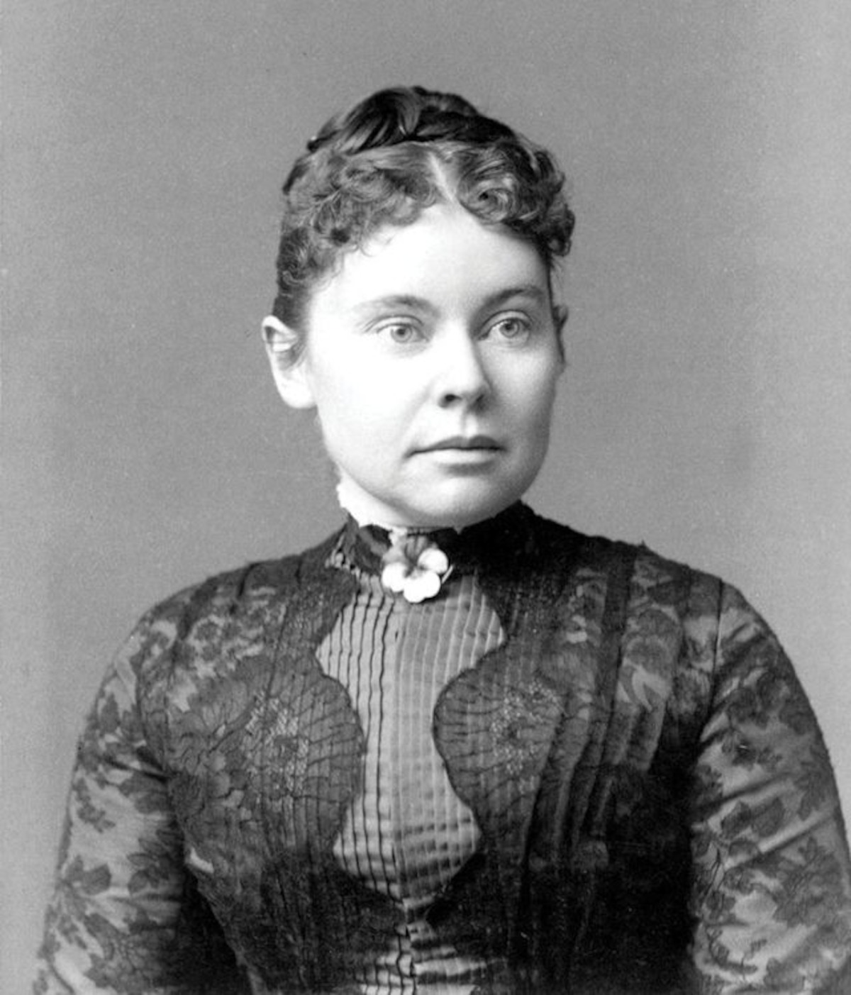 Woman in 19th-century black lace dress with floral brooch, neutral expression, posed against plain studio backdrop, formal historical portrait setting.