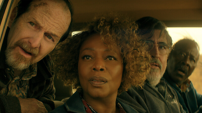 Four adults sit closely together inside a car, looking intently out the window. The lighting is warm, and the expressions on their faces suggest concern or curiosity. The car interior and a glimpse of the outdoors are visible.