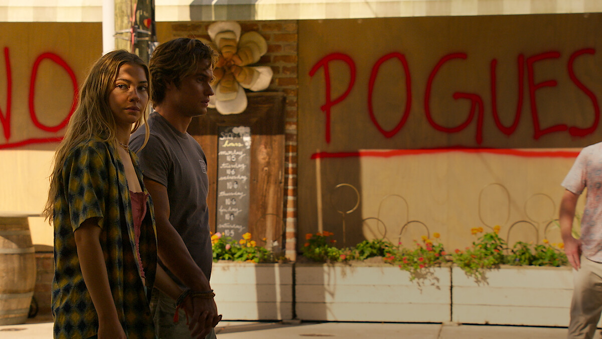 Two young people stand in an outdoor urban area with plants and a wall behind them featuring large red graffiti that reads "NO POGUES," creating a tense atmosphere.