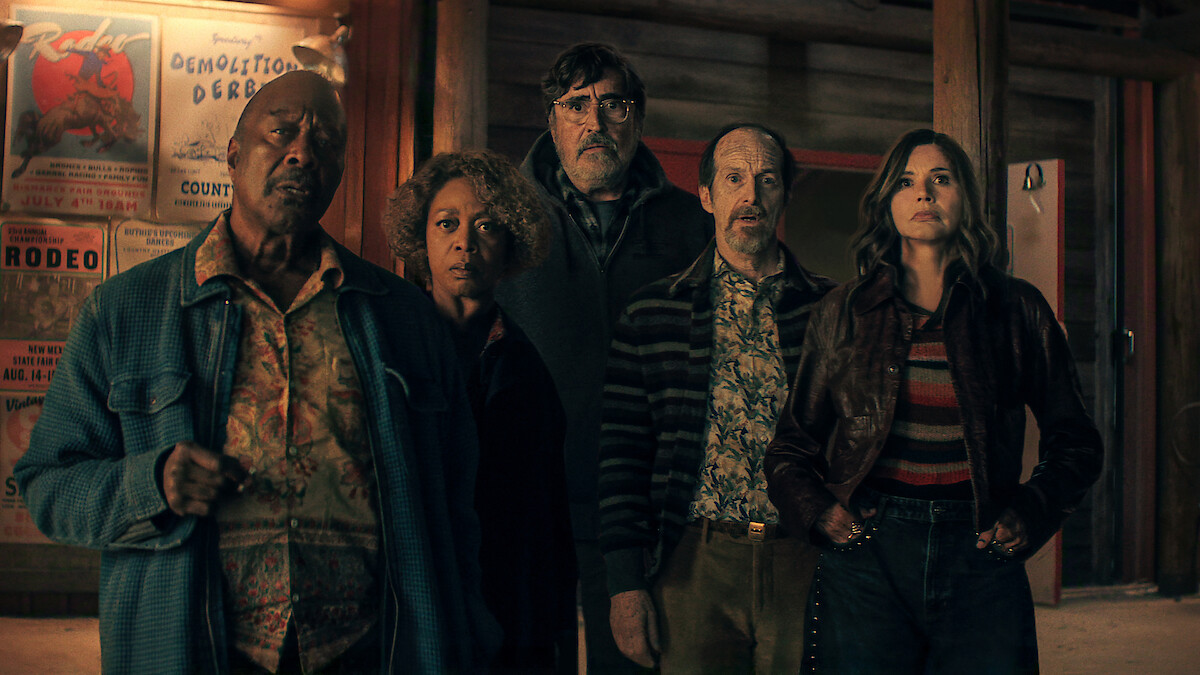 Five people stand indoors in front of wooden walls with posters, looking intently ahead, wearing retro-style clothing in a dimly lit setting suggestive of a bar or lodge.