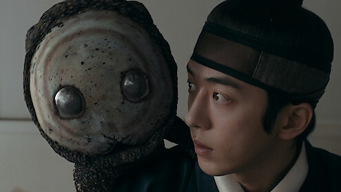 A young man in traditional Korean attire closely observes a large, round, alien-like puppet or creature with big metallic eyes in an indoor setting, suggesting a scene from a historical drama with fantasy elements.