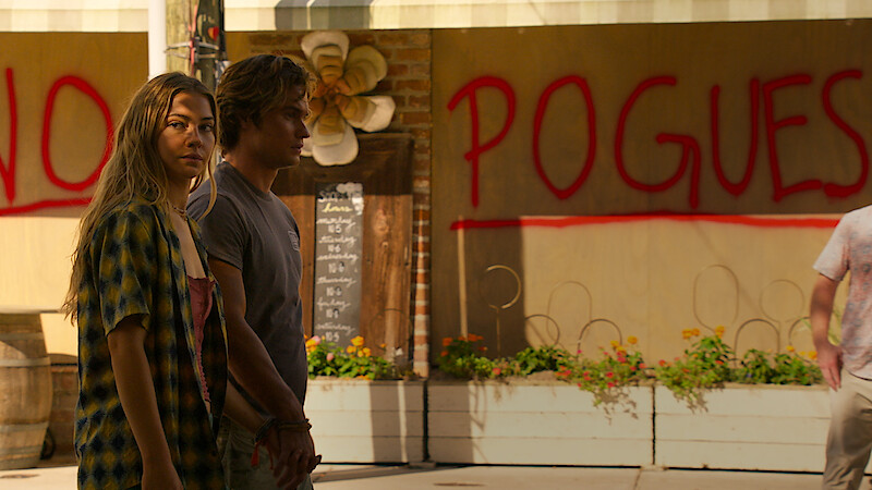 Two young people stand in an outdoor urban area with plants and a wall behind them featuring large red graffiti that reads "NO POGUES," creating a tense atmosphere.