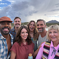 Grupo de seis adultos em roupas casuais, sorrindo para uma selfie diante de montanhas, deserto e um céu nublado. Eles têm uma aparência feliz e tranquila, estão vestindo roupas casuais e desfrutam da natureza.