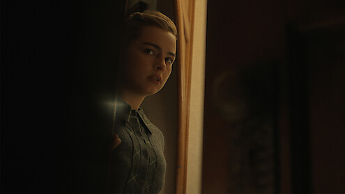 Addison Rae with a concerned expression peeks around a doorway into a dimly lit room.