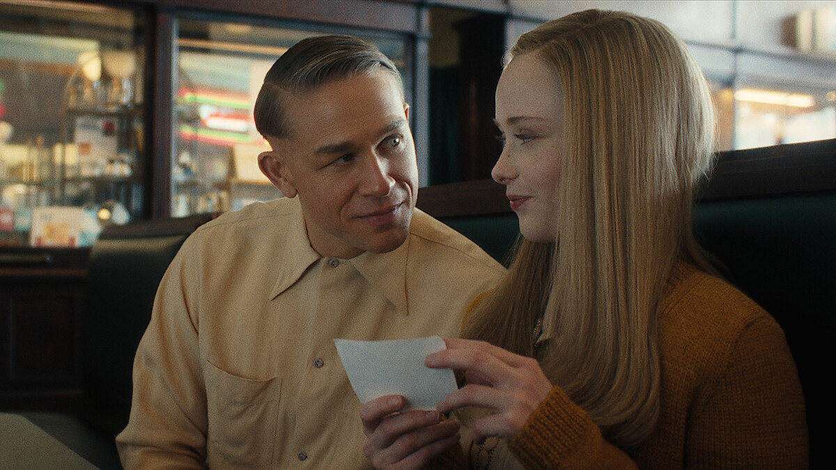 A man and woman, seated in a diner booth, share a sweet moment, smiling and holding a piece of paper between them.