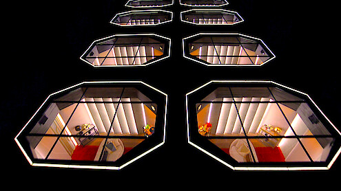Fileiras de janelas geométricas modernas e iluminadas na fachada escura de um prédio à noite, revelando interiores elegantes com detalhes em vermelho e laranja, cadeiras e pequenas mesas, que formam um padrão arquitetônico futurista.