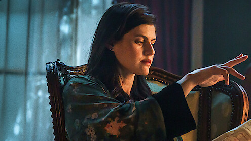 Woman sitting on ornate chair in dimly lit room, wearing a patterned robe, focused with her hand raised as if performing a gesture or casting a spell, with soft light coming through a window in the background.