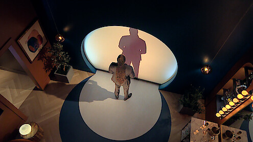 Dr Vic St John standing alone in a modern, dimly lit room with blue walls, casting a large shadow onto a circular white light on the wall. Decorative plants, artwork, and a set dining table create a stylish, intimate environment.