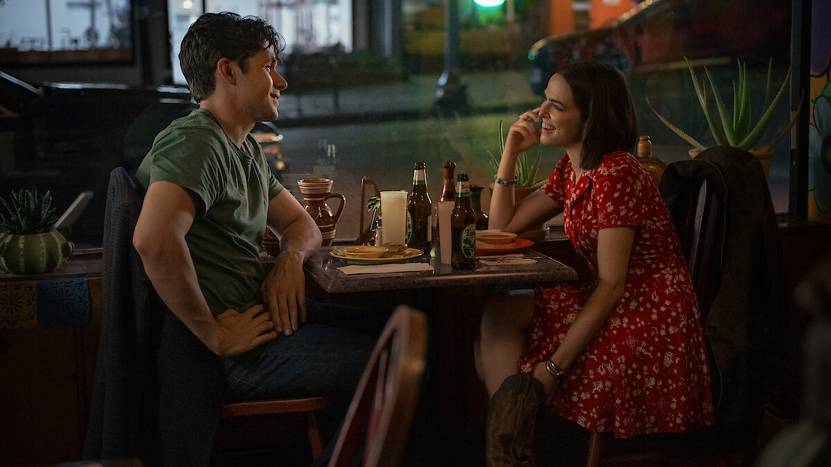 A man and woman sit across from each other at a cozy restaurant table at night, smiling and talking over food and drinks, with a street visible through the window behind them.