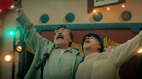 Two older men joyfully sing together on stage with arms raised, surrounded by stage lights and festive decorations in an indoor event setting, suggesting a celebratory or performance atmosphere.