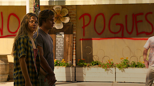 Two young people stand in an outdoor urban area with plants and a wall behind them featuring large red graffiti that reads "NO POGUES," creating a tense atmosphere.