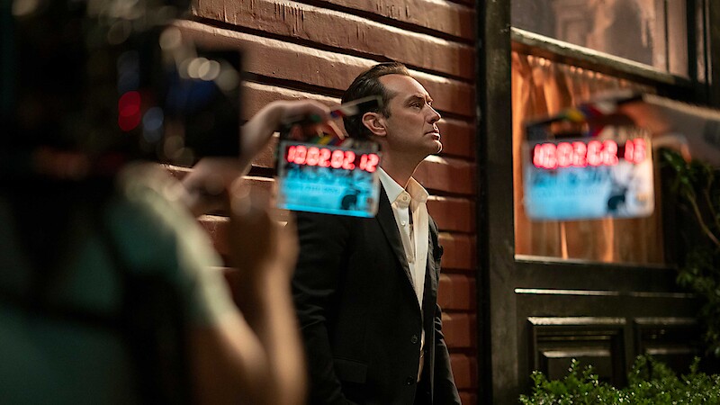 A man in a suit stands against a wooden wall at night, looking up thoughtfully. Movie cameras and crew are visible, suggesting a film set environment. Warm lighting from windows creates a cinematic atmosphere.