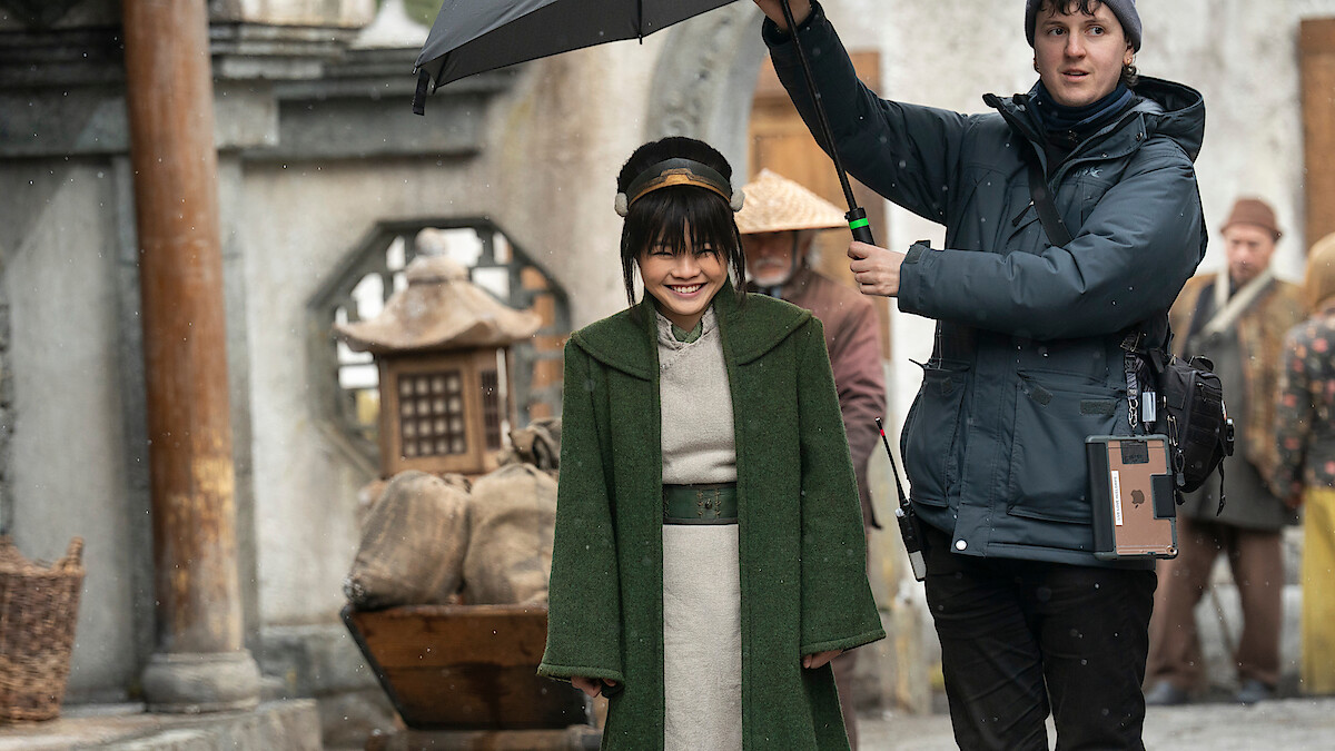 Miyako as Toph in a green coat stands outdoors on a film set while a crew member holds an umbrella over her, with people and rustic props in the background.