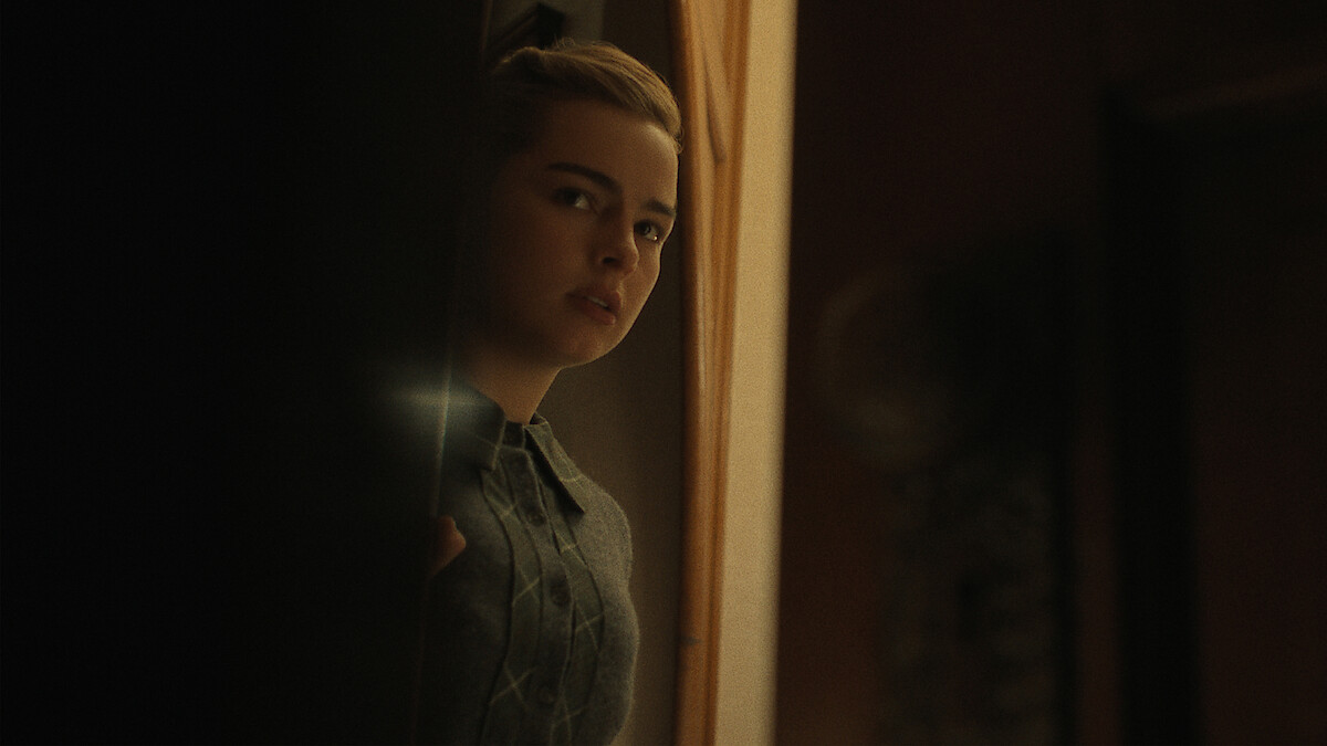 Addison Rae with a concerned expression peeks around a doorway into a dimly lit room.
