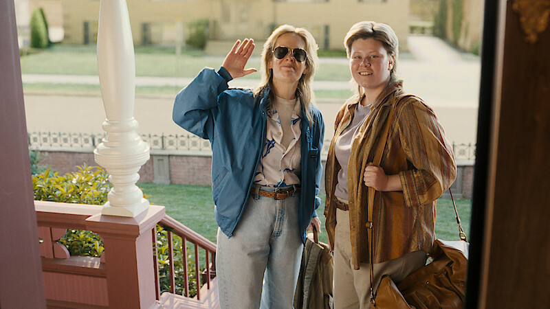 Two women with bags stand on a porch, one waving and the other smiling, both dressed casually with retro style; suburban houses and a lawn are visible in the background, suggesting arrival or greeting at a home.