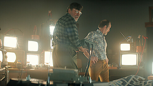 Two men in a dimly lit room filled with old televisions and electronic equipment, one holding a device, standing beside a bed with a patterned blanket, suggesting an experimental or scientific setting.