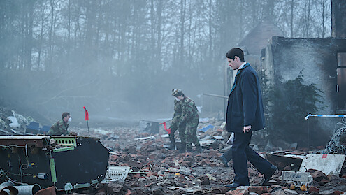 A man in a suit walks through a debris-strewn, foggy ruin as two people in camouflage uniforms investigate the wreckage; trees and a partially destroyed building are in the background.