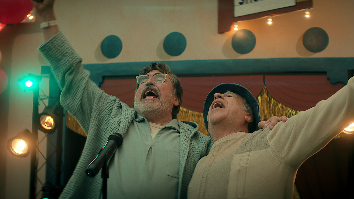 Two older men joyfully sing together on stage with arms raised, surrounded by stage lights and festive decorations in an indoor event setting, suggesting a celebratory or performance atmosphere.