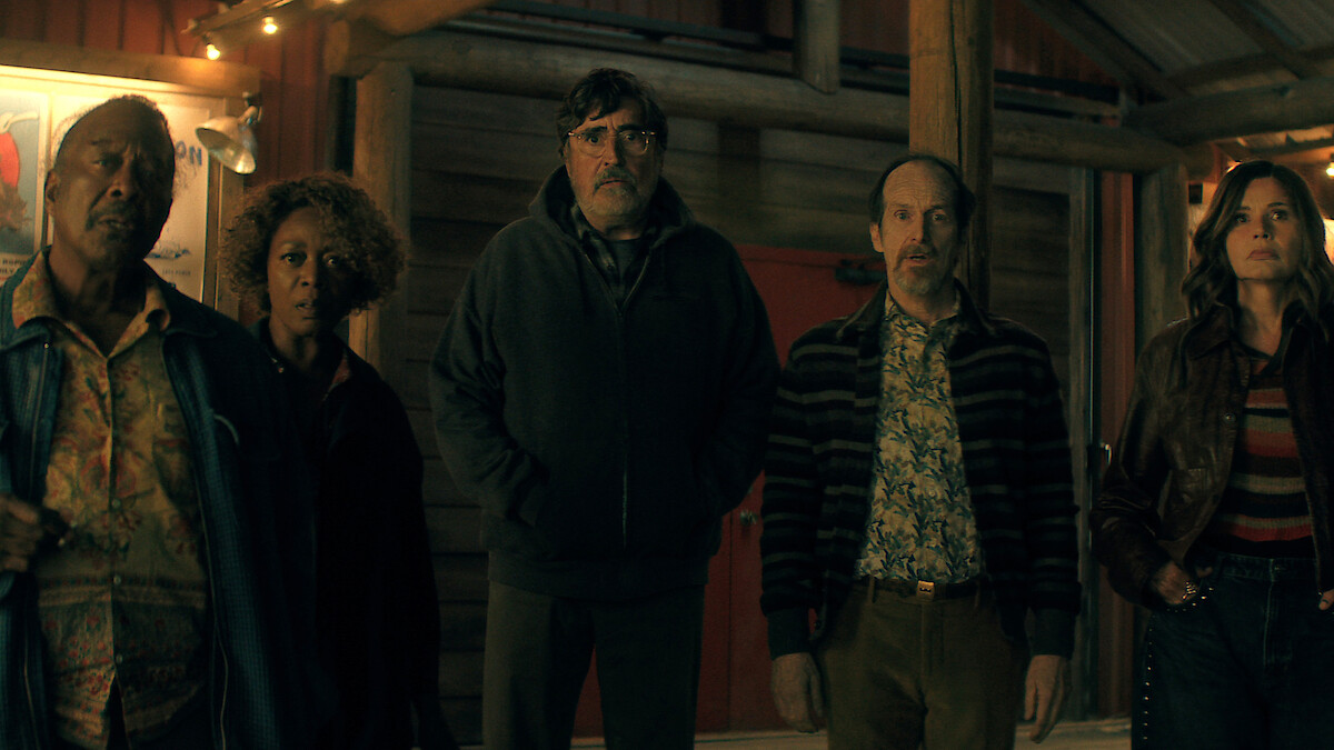 Five adults stand together outside a wooden cabin at night, dressed in casual clothes, looking serious or concerned, with warm string lights illuminating the rustic environment.