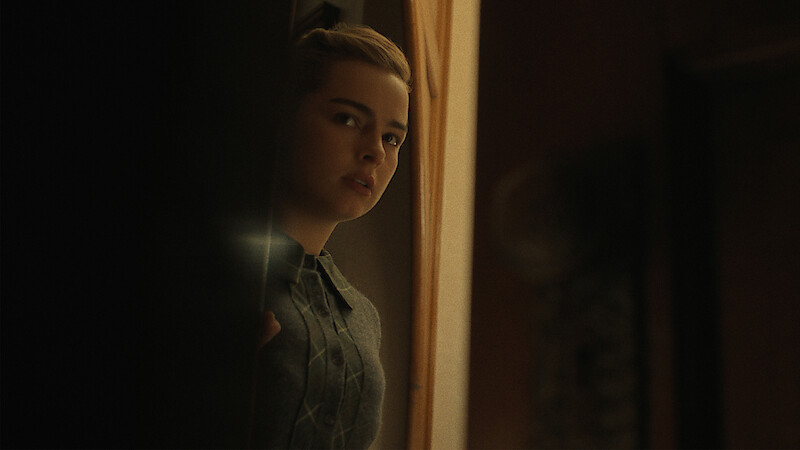 Addison Rae with a concerned expression peeks around a doorway into a dimly lit room.