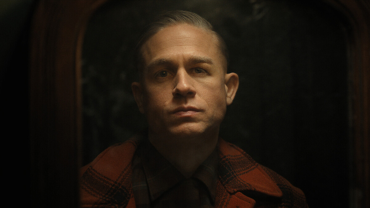 Ed Gein in a plaid jacket looks at his reflection in the mirror in a dark room.