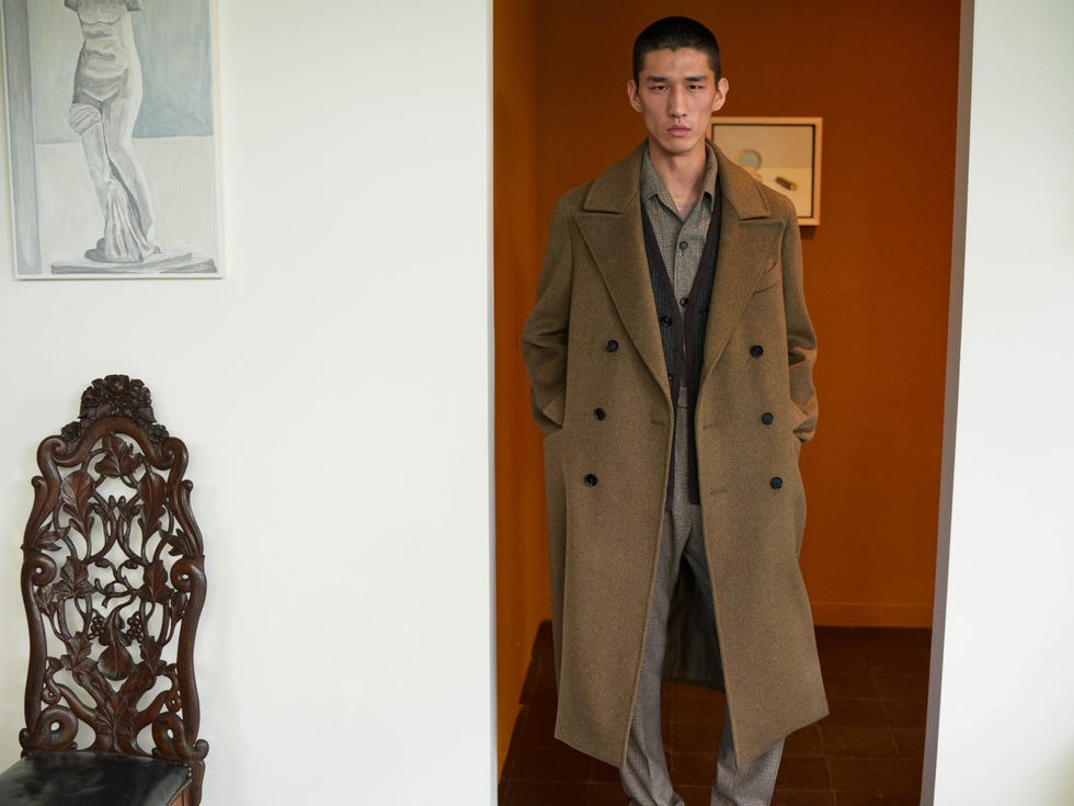 Loro Piana FW 2026/27 modello indossa un cappotto loro piana marrone sopra un completo sartoriale, in un interno elegante con opere d’arte e arredi d’epoca.