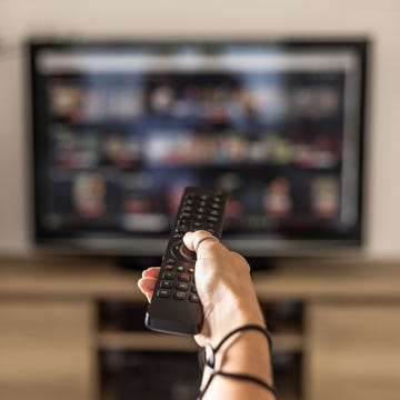 A woman is holding a remote control of a smart TV in his hand. In the background you can see the television screen with streaming entertainment apps for video on demand. woman watching tv