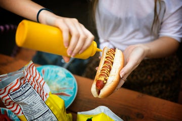 Cropped image of woman pouring mustard sauce on hot dog at garden party dietitians explain ultra-processed foods - Cropped image of woman pouring mustard sauce on hot dog at garden party