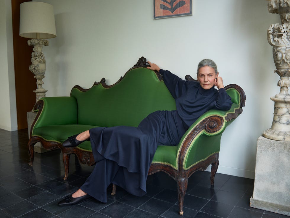 Loro Piana FW 2026/27 donna indossa un elegante abito blu navy loro piana, seduta su un divano vintage verde in un interno raffinato.