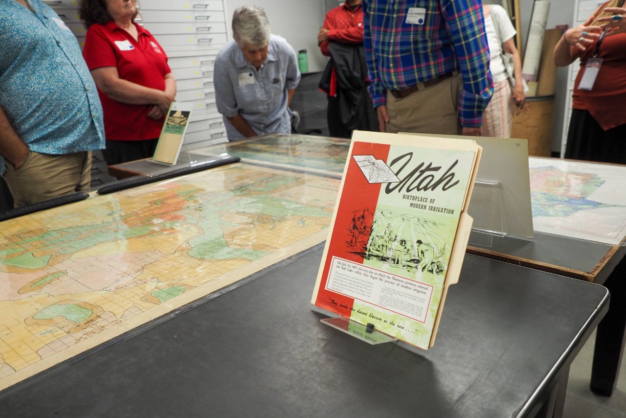 People stand around tables displaying maps and a book titled Utah in a library or archive.