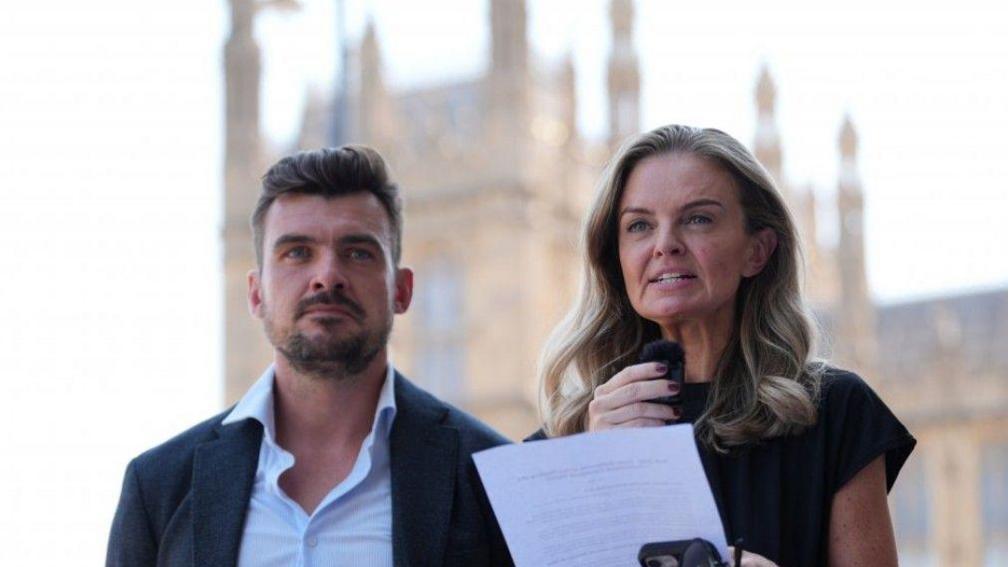Laura Enston (centre), and Stephen Beard, the grandchildren of Ruth Ellis, making a statement to media outside the Houses of Parliament. Enston has shoulder length, dark blonde hair and wears a short-sleeved black dress with purple nail varnish. She is holding a piece of paper with her statement written on it.