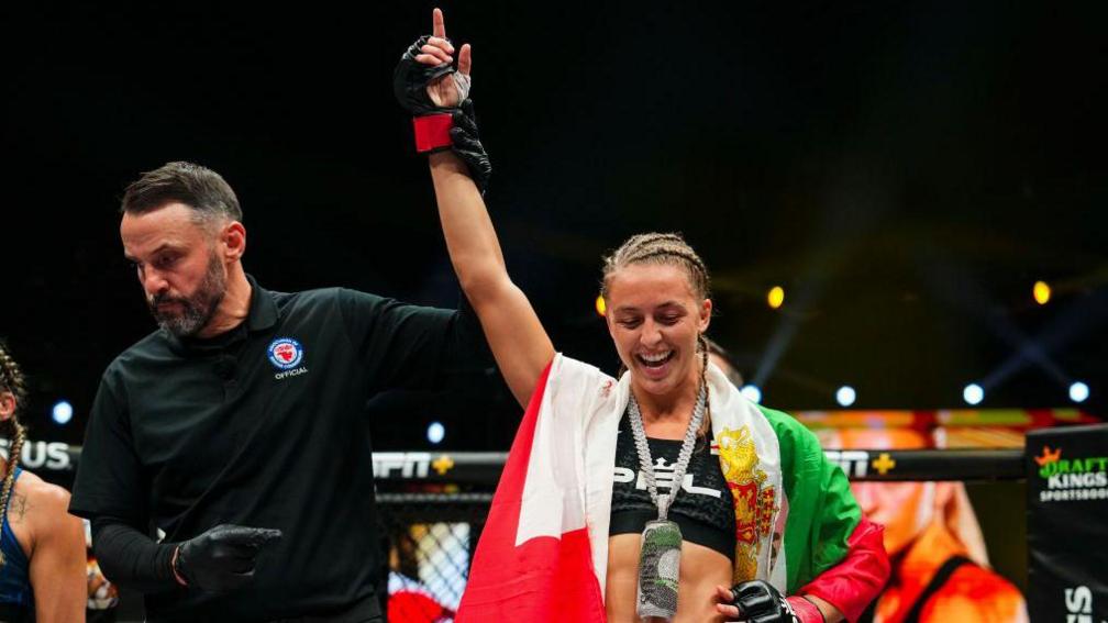 Dakota Ditcheva has a flag on her shoulders and her arm is being raised in the air