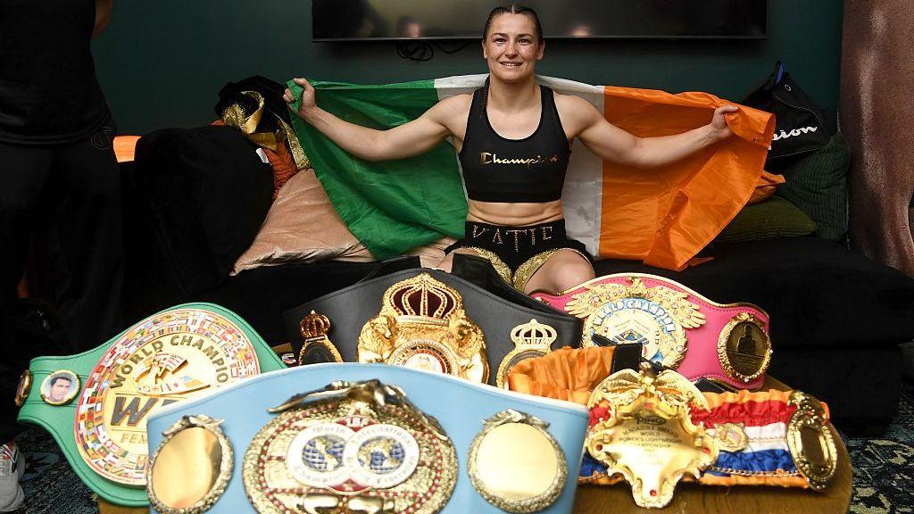 Katie Taylor holds up the Republic of Ireland flag and is sat behind a table with her light-welterweight world titles on top