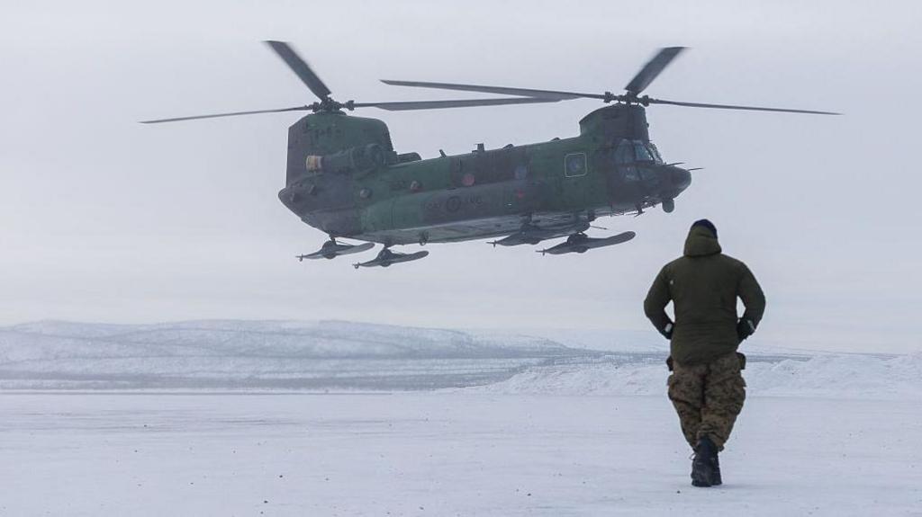 Canadian soldier walking towards a green Chinook helicopter equipped with skis in a frozen plain.