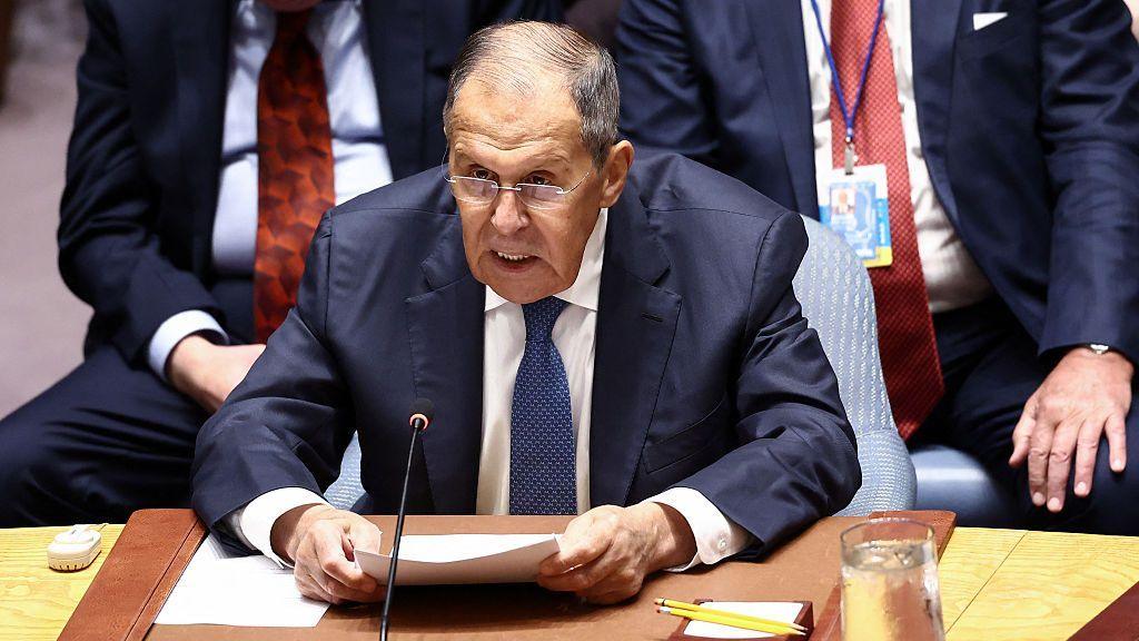 Sergei Lavrov wearing a navy suit, white shirt, and blue tie. He is sat behind a microphone with other diplomats in suits behind him. Lavrov is holding several documents in his hands, wears glasses, and has short grey hair.