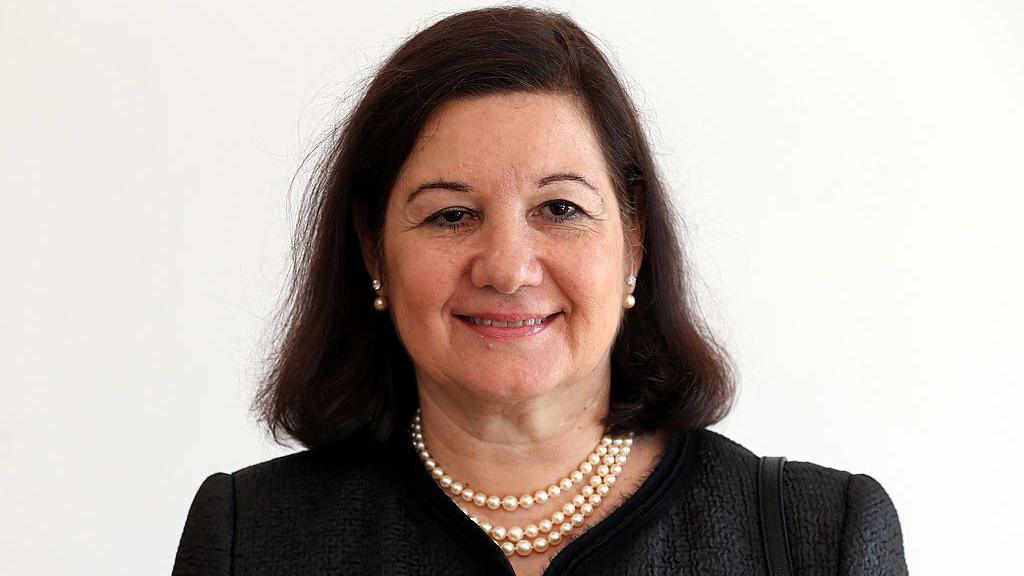 A head and shoulders shot of a woman with dark, shoulder-length hair wearing a dark top and a pearl necklace
