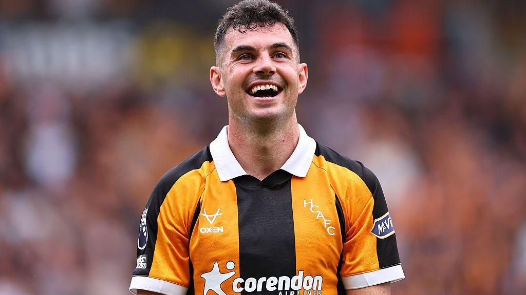 John Egan, in Hull's black-and-amber-striped kit, smiles while looking at the crowd during a Premier League match