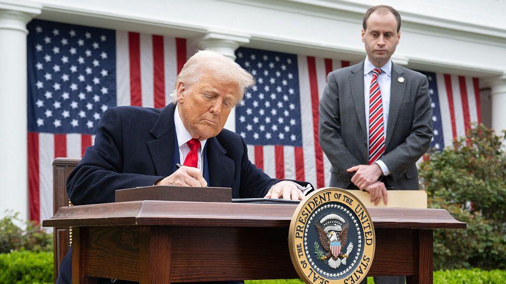 Donald Trump sits at a desk in the White House rose garden and signs an executive order. American flags hang behind him as an aide waits to the side.