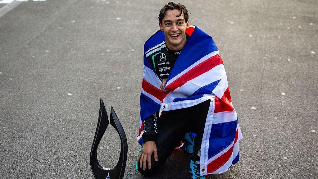 George Russell wrapped in the Union Jack flag, posing with the Azerbaijan Grand Prix trophy