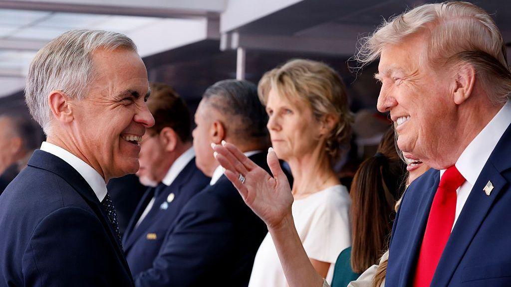 Mark Carney and Donald Trump, each in profile, share a smile . A crowd can be seen in the background.