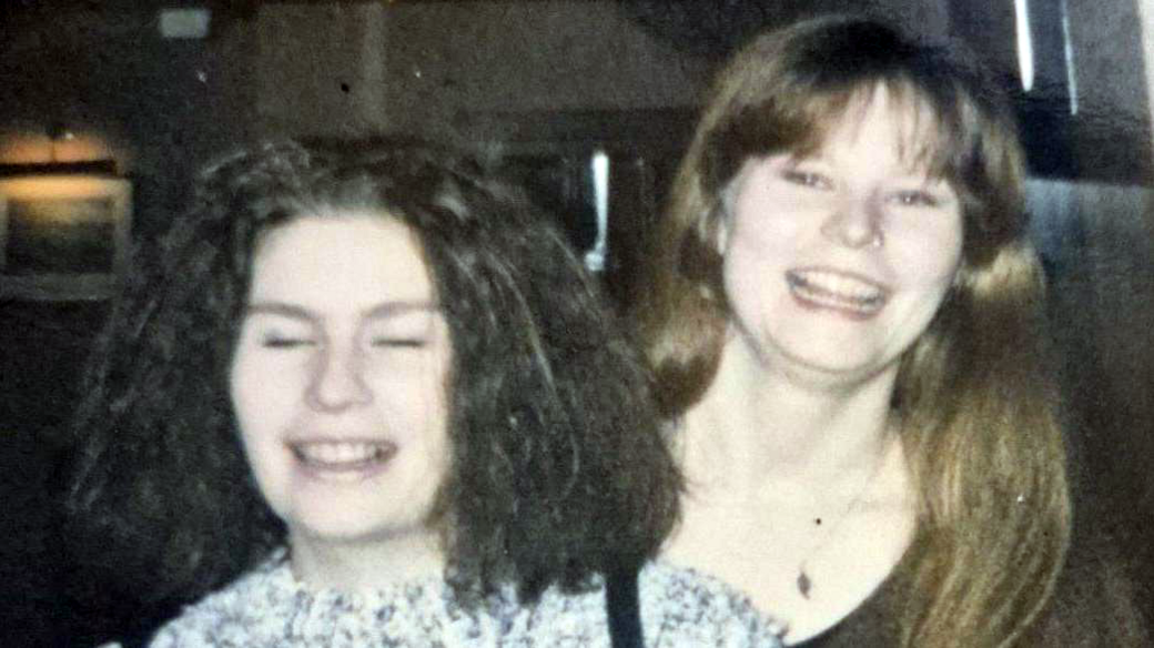 An old photo of two young women on a night out. They are grinning at the camera and standing beside each other. One has dark hair and the other has brown hair. They are standing in a darkly lit indoor venue.