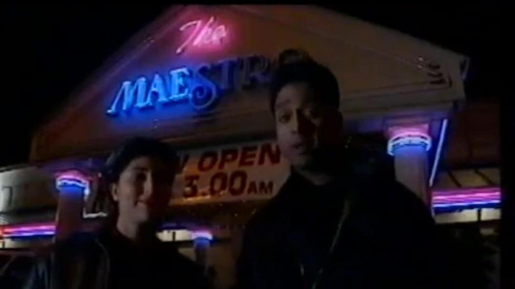 Two young South Asian people standing in front of a building with a neon pink and blue sign saying 'The Maestro', and 'open until 3am'