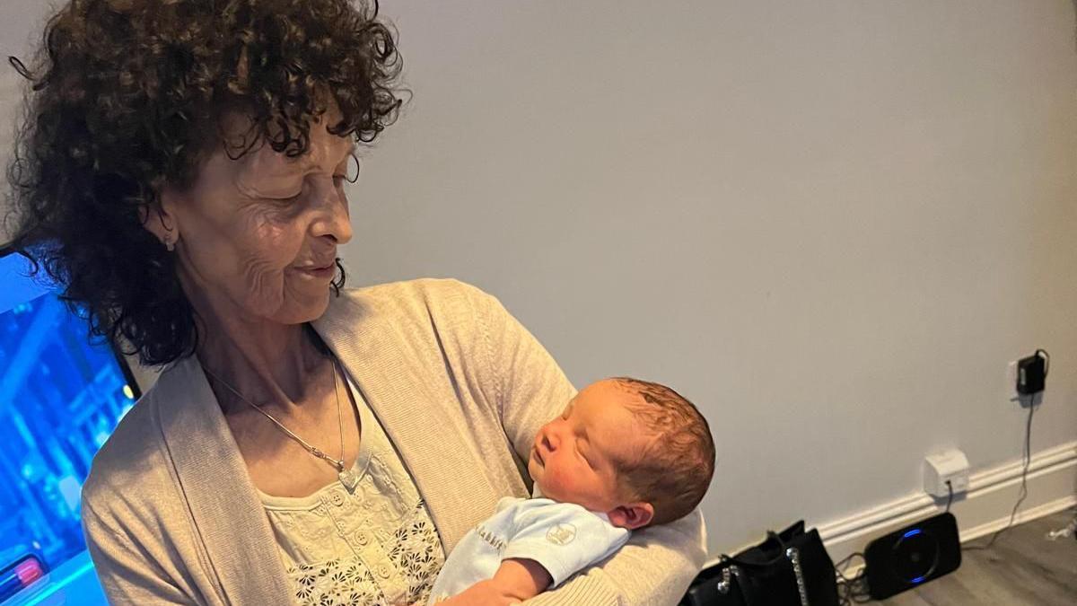 An older woman with curly dark hair smiles down at a newborn baby she is holding.