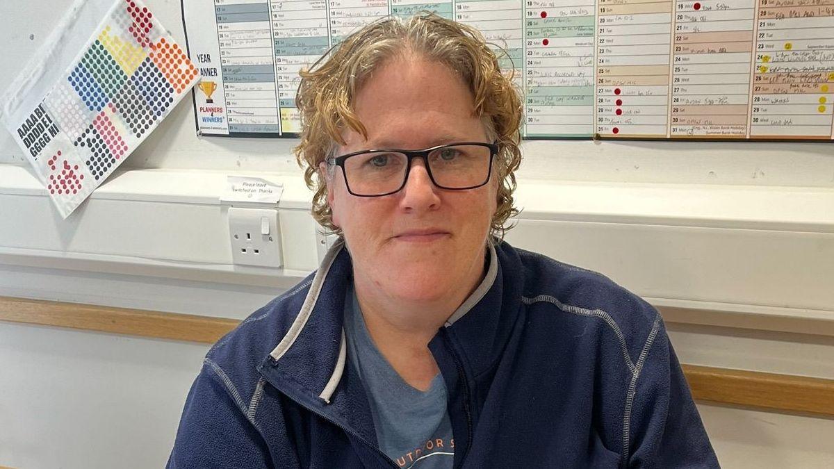 Trudi, a woman with curly blonde hair and glasses, sits at a desk in a blue zip-top with a blue T-shirt underneath. There is a wall planner behind her and plug points.