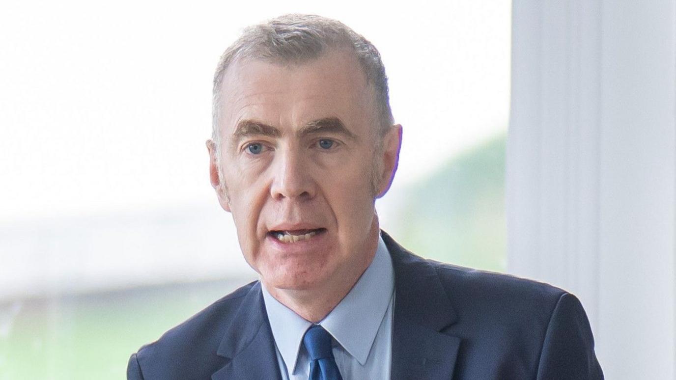 Adam Price speaking at an event in front of a window, wearing a blue suit, blue shirt and a blue tie.